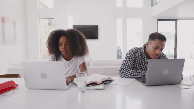 Black teenage boy helping his sister while they sit at home using laptop computers, front view